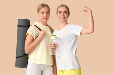 Beautiful young sporty sisters with yoga mat and bottle of water showing muscles on beige background