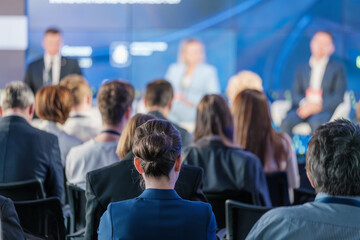 Audience members attending a business conference with blurred speakers on stage discussing important topics.