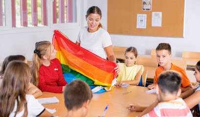 Schoolchildren sitting in classroom and listening to female teacher. She holding rainbow flag in hands and talking about minorities