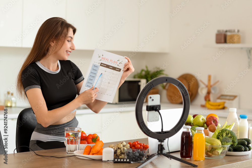 Wall mural sporty young woman with meal plan and healthy food recording video in kitchen. diet concept