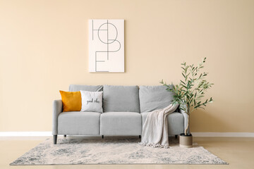 Soft sofa with plaid and cushion, picture on wall and decorative olive tree in interior of living...