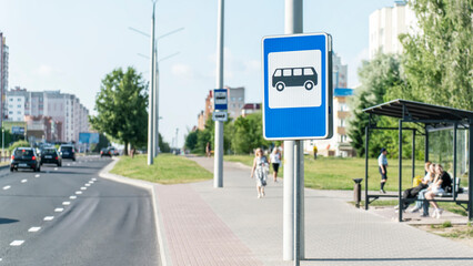 Blue bus stop sign stands prominently along city street with bus shelter and waiting passengers...