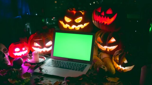 Jack O Lanterns and laptop with green screen, place for ad offers, next to Halloween carved pumpkins and flashing light, candles, colorful garlands, night party background.