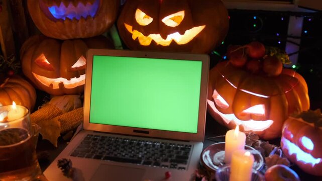 Laptop with chromakey and traditional Halloween attributes carved pumpkins and burning candles, mug beer, saints night party background.