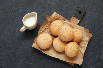 Wooden board of fresh buns with sesame seeds and jug of milk on dark background