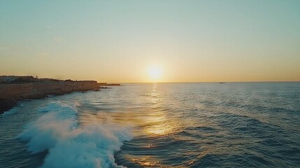Fototapeta premium The sun sets over the ocean as a wave crashes on the shore and buildings stand tall on the opposite side of the water