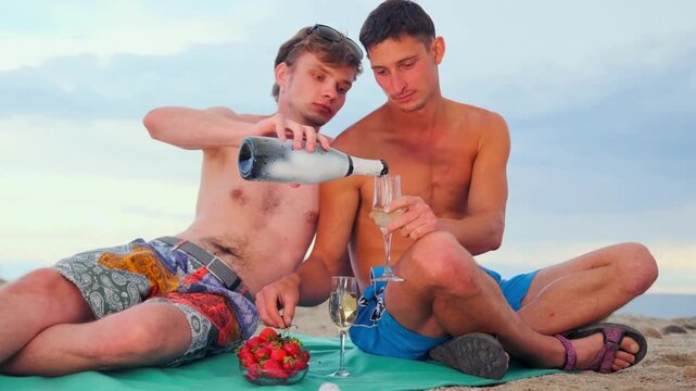Two shirtless gays on a romantic date drinking wine pouring it into glasses sitting on picnic mat with strawberries on beach. Happiness, freedom and love concept.