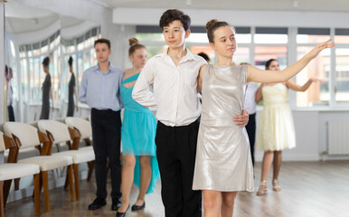 Fototapeta premium Happy interested teenage students preparing for college festive event, rehearsing ballroom dance steps in spacious classroom while female teacher observing from background..