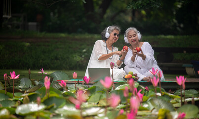 two elderly women sitting by a pond surrounded by blooming lotus flowers. They are both wearing white tops and headphones, enjoying music while holding apples