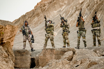 A group of military men in combat gear patrol in the middle of a desert and tropical jungle....