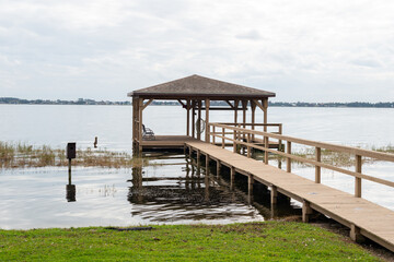 A wooden pier with a large square hut at a wharf. The platform marina has a wooden rail along the bridge and a metal roof. The gazebo stage is over a blue calm ocean. The sky is blue with some cloud
