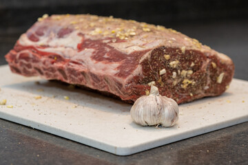 A large rack of raw beef prime rib on a white industrial plastic cutting board. The meat is sitting on the kitchen counter next to a large bulb of garlic. The roast is marinated in minced garlic.