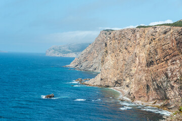 A rugged coastline and cliff in the foreground near a deep blue ocean. It's a bright sunny day with a blue sky and few clouds. There's an island in the background with greenery, buildings, and rock. 