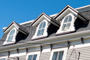 Three peaked dormer-style windows on a hip roof covered with black shingles. The vintage white wooden frames have arch-shaped glass and closed single-hung panes with white curtains hanging inside.  