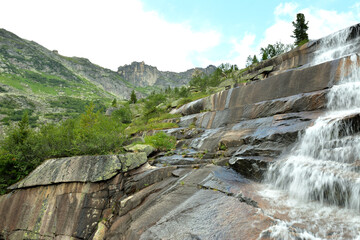 Steep stone steps from which a rapid waterfall cascades down in a coniferous forest under a summer cloudy sky.