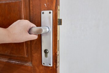 Human hand with light skin tone with opening door gesture isolated on horizontal brown wooden door with metal knob background.