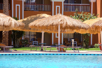 Empty deck chairs under straw shade umbrellas on swimming pool side in tropical resort. Summer vacations and getaway concept