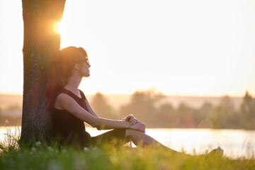 Lonely young woman sitting alone on grass lawn on lake shore enjoying warm evening. Wellbeing and relaxing in nature concept