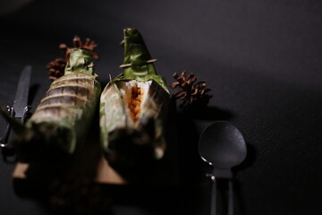 Grilled rice wrapped in banana leaf, served on brown burlap fabric with a black background, evoking a rustic, traditional vibe.