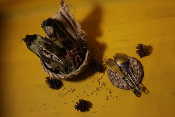 Traditional food wrapped in banana leaves placed in a woven basket, with seeds scattered around on a yellow background.