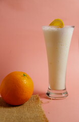 Minuman susu dingin dengan irisan jeruk dan satu butir jeruk, disajikan dalam gelas dengan latar belakang merah muda cerah, menyegarkan dan manis.