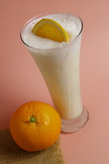 Minuman susu dingin dengan irisan jeruk dan satu butir jeruk, disajikan dalam gelas dengan latar belakang merah muda cerah, menyegarkan dan manis.