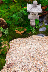 Minimal Japanese garden in home with small stone lantern and bamboo fountain and moss and flowers.
