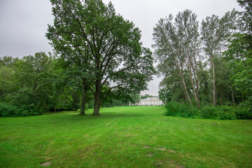 A vibrant green meadow stretches out under a cloudy sky, framed by tall trees and leading to a distant historic estate, evoking tranquility and natural beauty.