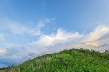 A vibrant blue summer sky, adorned with fluffy white clouds, stretches above a lush green hilltop. The grass sways gently in the breeze, creating a tranquil and peaceful scene.