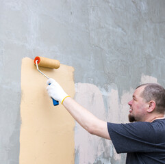 A man wearing gloves paints a wall with a roller brush. The wall is gray, and he is applying a coat of tan paint. The sun casts a warm glow on the wall and the man's face.