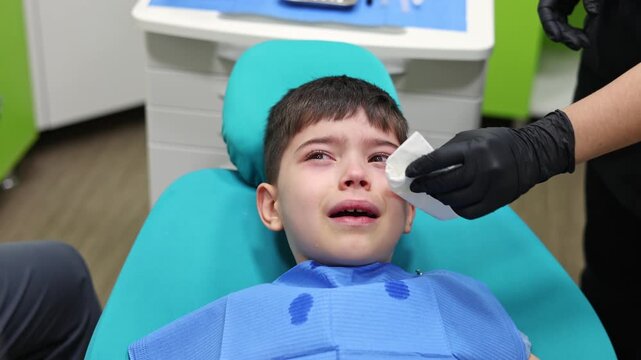 kid in dental office. dentist checking child teeth using tools mouth mirror and scalar.crying with tears, sad boy is afraid.doctor looking at screen,tooth filling treatment.kid in chair being examined