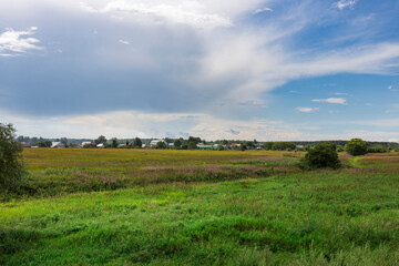 Obraz premium Lush green fields stretch towards a small town, illuminated by the golden hues of the late afternoon sun against a backdrop of soft, fluffy clouds.