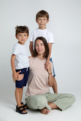 Full length image of a sons hugs his mother, they are sitting together on the floor, looking at camera, over grey background.