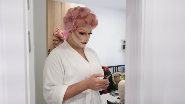 A drag queen wearing a bathrobe and curlers looks at their smartphone while getting ready at home for a performance.