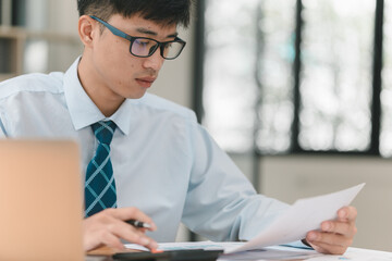 A man wearing glasses and a blue shirt is sitting at a desk with a piece of paper in front of him. He is looking at the paper and he is focused on it