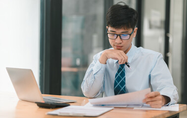 Fototapeta premium Focused Determination: A young businessman in a crisp blue shirt and tie, glasses perched on his nose, meticulously reviews paperwork at his desk, a laptop and calculator nearby. His focused expressio