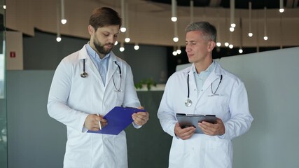 Two male doctors wearing lab coats discuss patient notes while standing in a modern hospital. One doctor writes on a clipboard. - Powered by Adobe