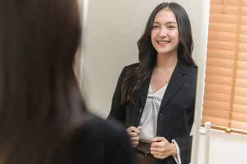 Young beautiful Asian woman in formal suit checking dressing tidiness and encourage herself in mirror at home before go for job interview. Woman recruitment employee or staff in company concept.
