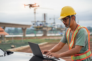 Asian male engineer using laptop and drone to inspect construction at bridge construction site. Construction area of ​​large bridge connecting road across river.