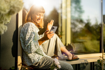 A woman relaxes by a window in a sunlit garden, checking her phone. Surrounded by blooming flowers, she enjoys a moment of solitude and peace, escaping to nature