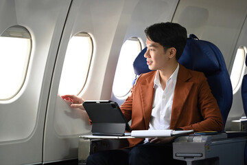 Handsome young businessman in a suit sitting in an airplane seat with a tablet in front of him