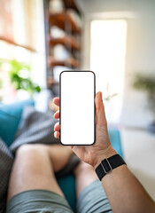 Man holding a smartphone with a blank screen in a home interior, sitting on a couch. Perfect for showcasing mobile apps or digital content. First-person perspective photo.