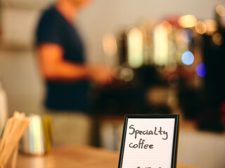 This image captures a small sign advertising specialty coffee with a blurred barista working in the background, highlighting a vibrant and active café ambiance.