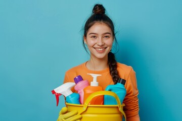 Joyful young housemaid in 20s with yellow gloves holding cleaning supplies bucket on blue background