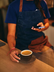 A barista in a blue apron holds a cup of coffee while checking a smartphone, emphasizing multitasking and the integration of technology in daily coffee shop routines.