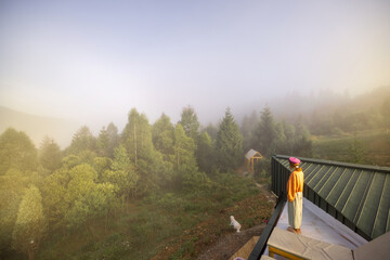 A woman stands on a rooftop, overlooking a misty forest and a small greenhouse, with her dog patiently waiting below, capturing the tranquility of a foggy morning in a secluded natural setting