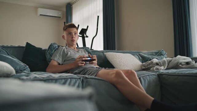 Focused teen boy sits on sofa in living room, plays online video game on console using wireless controller. Caucasian teenager spending leisure time on weekend. Lifestyle concept. Gaming at home.
