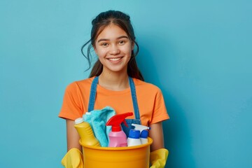 Happy young maid in twenties with yellow gloves holding cleaning bucket on blue background