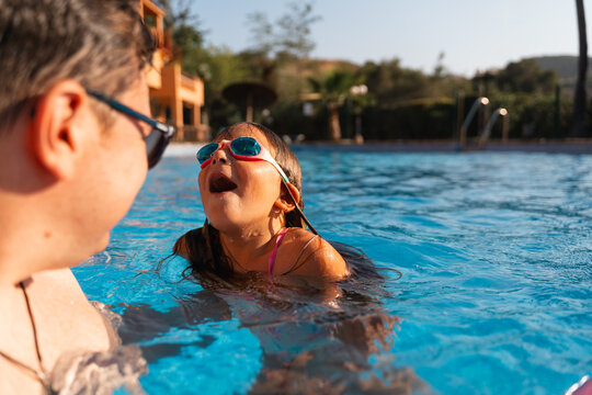 A young girl with goggles looks up excitedly while swimming with her father in an outdoor pool, enjoying a sunny day together