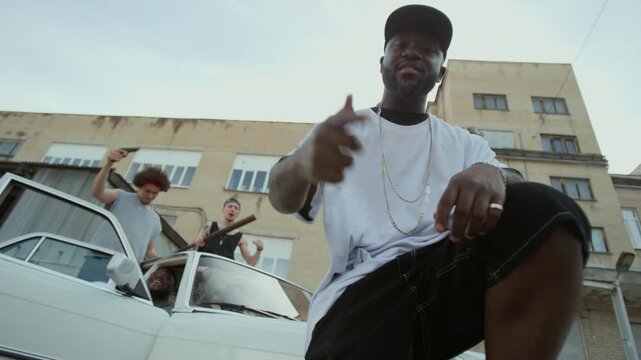 Low angle shot of cool Black male singer in streetwear and silver necklace pointing at his friends performing behind in car and rapping at camera outdoors in back alley